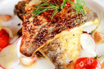Grilled Red Snapper on a white plate, garnished with dill. Around the plate are slices of grilled onions and grilled cherry tomatoes.