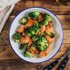 Cooked cubes of chicken and broccoli florets in a white bowl with blue trim on a wood table.