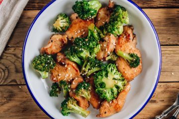 Cooked cubes of chicken and broccoli florets in a white bowl with blue trim on a wood table.