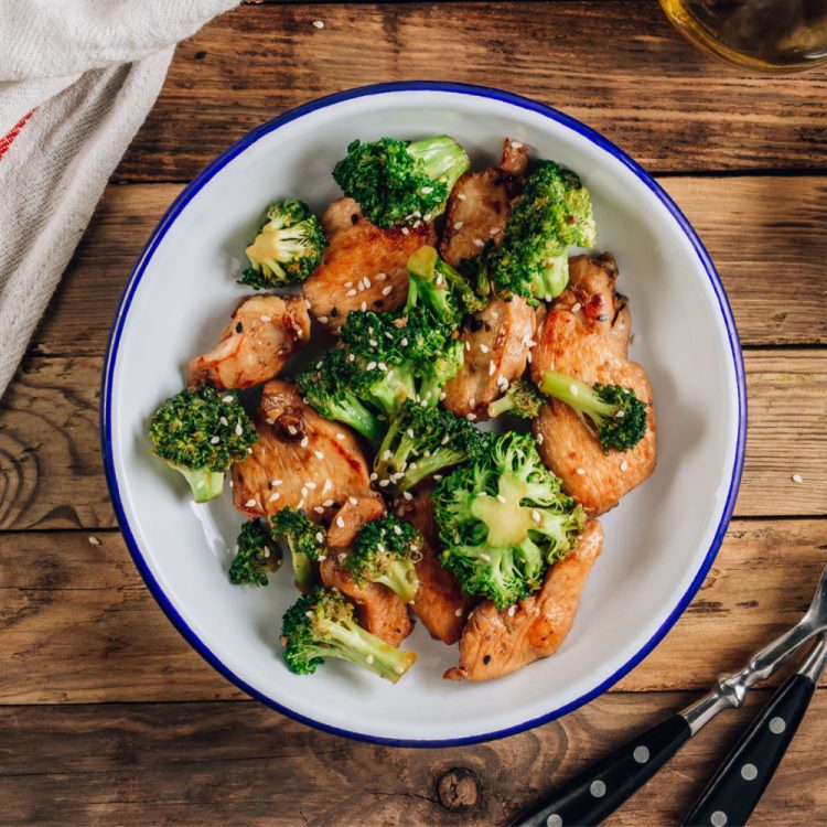 Cooked cubes of chicken and broccoli florets in a white bowl with blue trim on a wood table.